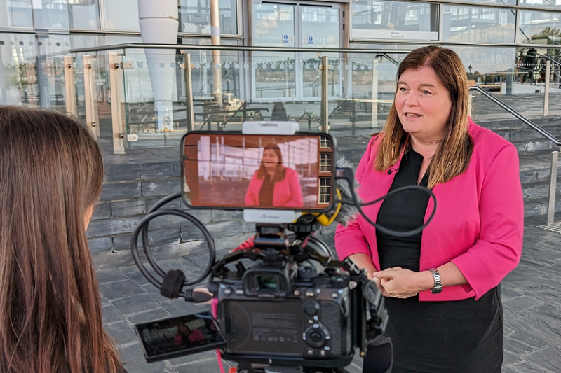 Sioned Williams MS is being interviewed for television and a camera can be seen in the foreground, with a small screen showing the shot of Sioned. Sioned Williams MS is standing on the Senedd steps, outside the Senedd building, the glass front of which is visible behind her.