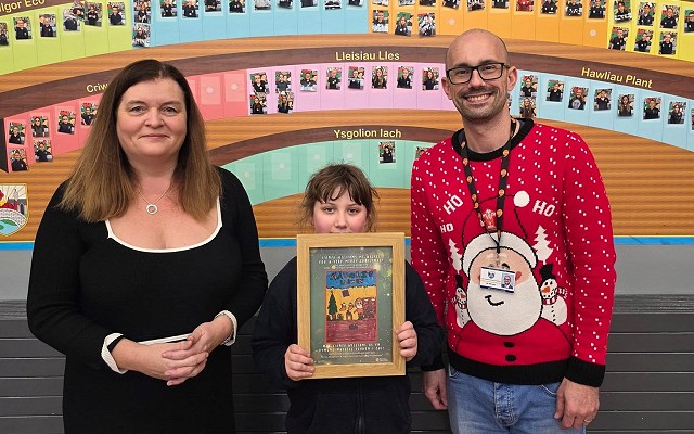 Sioned Williams stands with Seren, and her teacher. Seren is holding a frame with a copy of her winning design in it. Behind them is a display wall with the words 