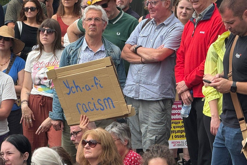 Llun o bobl yn y rali Stand Up To Racism yn Abertawe. Yng nghanol y llun mae arwydd cardbord sy'n dweud “Ych-a-fi Racism” (ffrwg, hiliaeth)