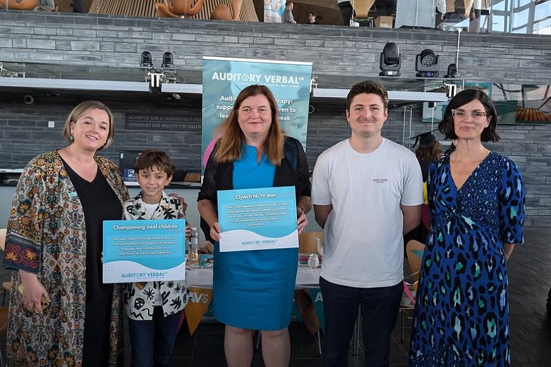 Llun grŵp yn y Senedd gyda Sioned Williams a gweithredwyr yn dal arwyddion Auditory Verbal UK yn hyrwyddo therapi lleferydd i blant byddar.