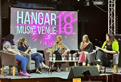 Panel discussion with five women seated on stage at Hangar 18 Music Venue, with a large screen displaying the venue name in the background.
