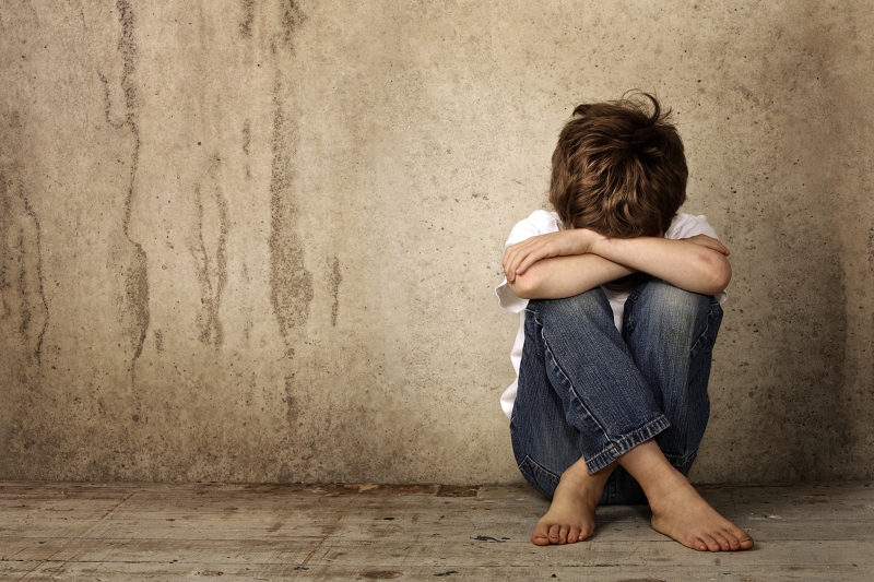 Boy in jeans and white t-shirt, no shoes, face hidden sitting cross legged against a stone wall background