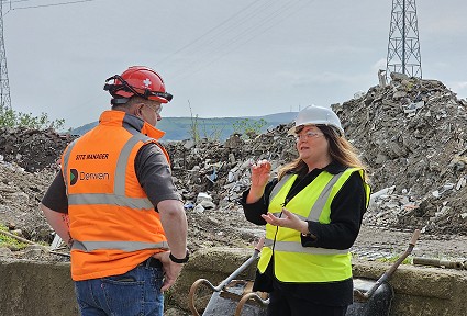 Sioned and a man both in hi-vis jackets and hard hats speaking in front of a large pile of rubbish at the recycling plant