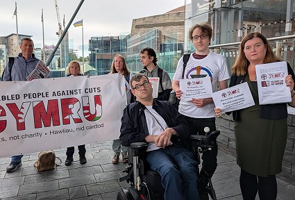 Sioned ac ymgyrchwyr y tu allan i'r Senedd yn dal baneri mae un mewn cadair olwyn