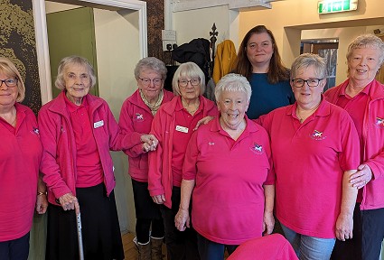 A group of older women wearing matching pink tops pose together indoors with Sioned Williams standing behind them
