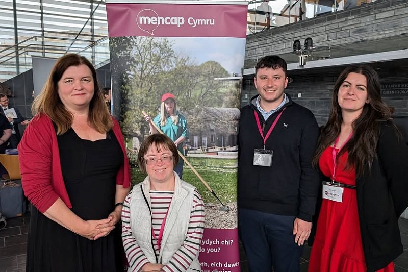 Llun grŵp o tri o bobl a Sioned Williams yn sefyll o flaen baner Mencap Cymru yn ystod Wythnos Anabledd Dysgu, wedi'i dynnu yn y Senedd.