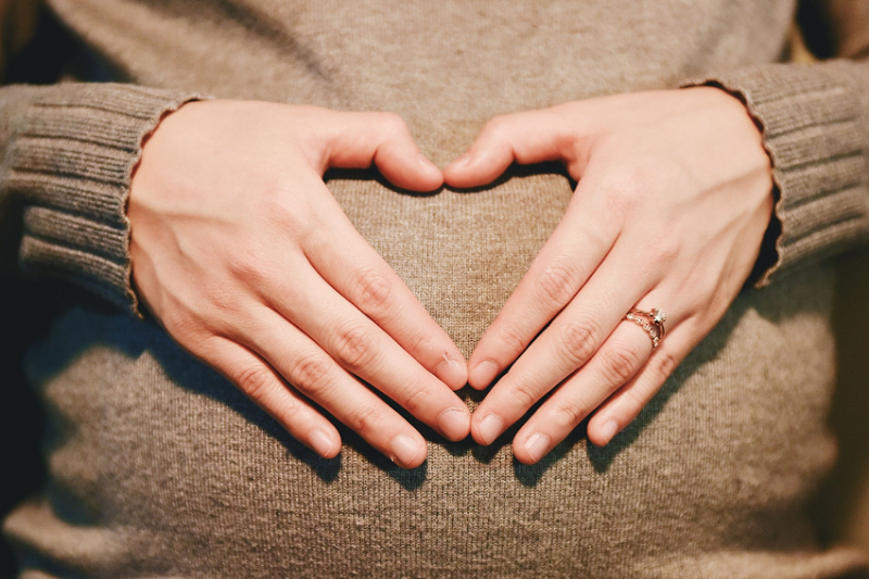 A lady touching the pregnant belly with two hands making the shape of a heart