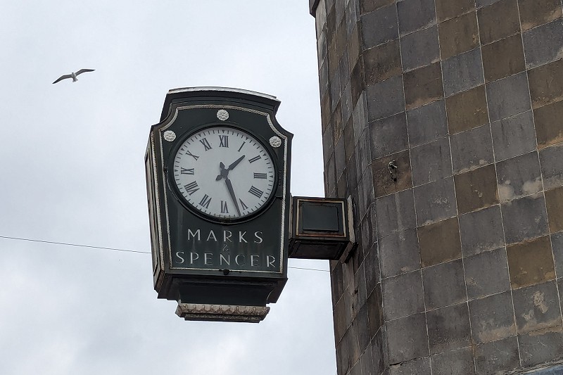 Marks spencer outside wall clock telling the time of 1:27pm with a view of a seagull flying behind