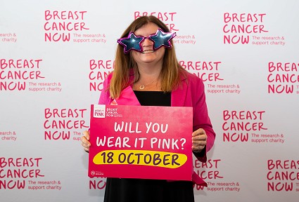 Sioned Williams MS wearing pink and holding a sign for Breast Cancer Now’s ‘Wear It Pink’ campaign, with star-shaped glasses and a backdrop promoting the charity