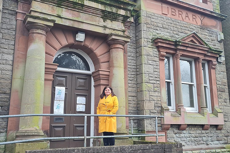 Sioned Williams yn gwisgo cot felen yn sefyll y tu allan i adeilad
