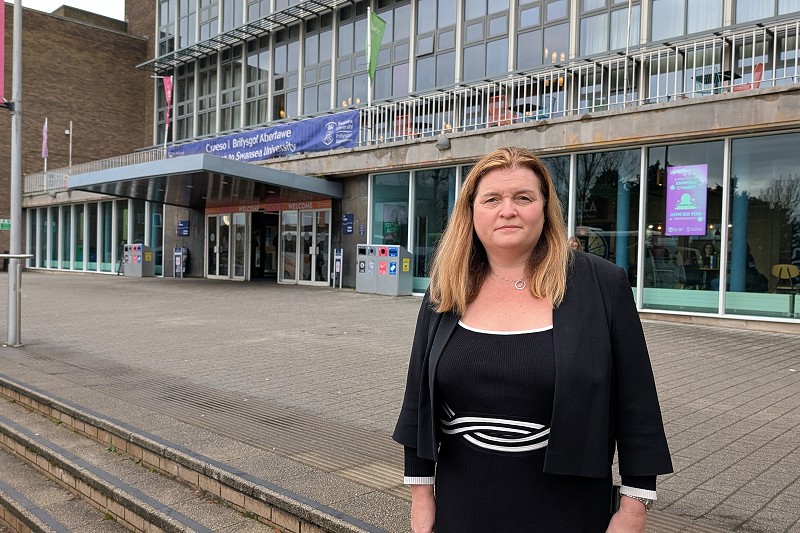 Sioned yn sefyll o flaen adeilad ar gampws Singleton Prifysgol Abertawe
