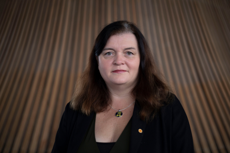 Sioned yn gwisgo du yn erbyn cefndir pren