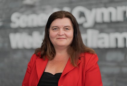 Sioned Williams AS yn edrych yn syth i'r camera o flaen adeilad Senedd Cymru yn gwisgo siaced goch