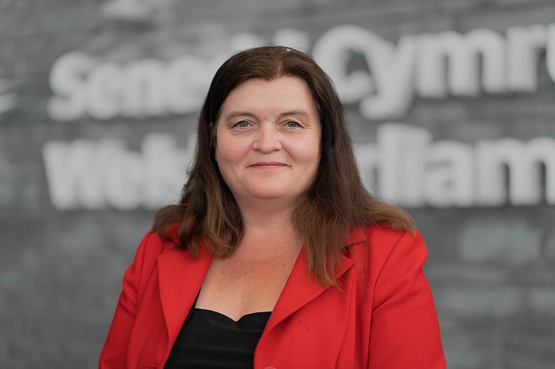 Sioned Williams AS yn edrych yn syth i'r camera o flaen adeilad Senedd Cymru yn gwisgo siaced goch