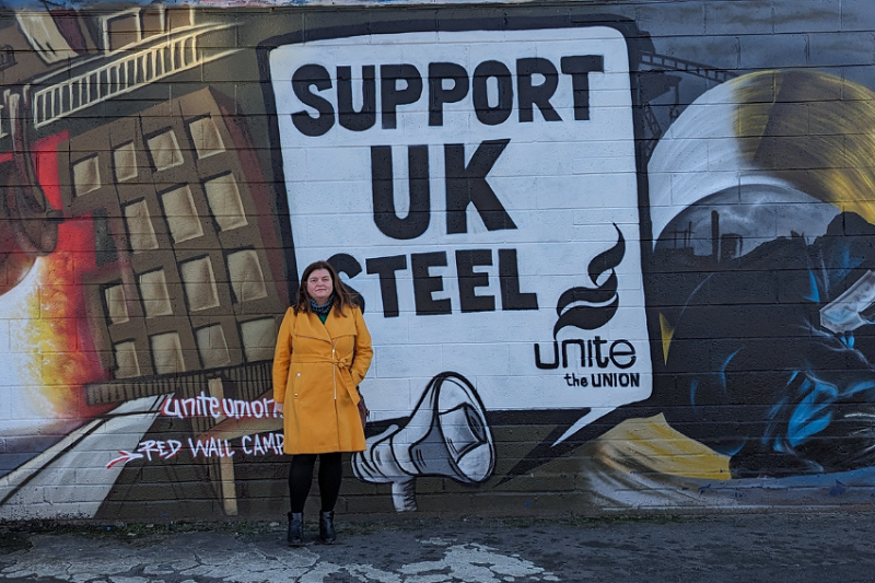 Sioned William yn sefyll o flaen celf wal graffiti Support UK Steel