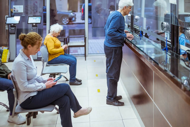 A view of a hospital reception area, with a gentleman standing at the reception desk and others seated, looking at their phones.